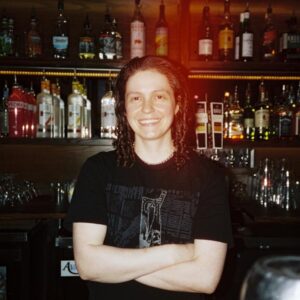 Josie, smiling with arms crossed, stands in front of a bar shelf with various liquor bottles.