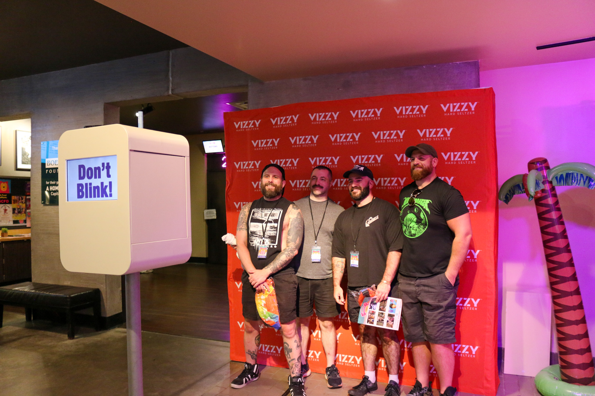 Four men smiling in front of Vizzy Hard Seltzer backdrop, adjacent to a sign reading "Don't Blink"