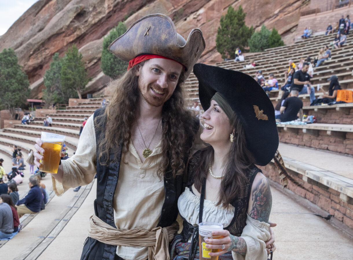 Man and woman dressed as pirates smiling, holding drinks in an outdoor amphitheater with rocky backdrop and trees