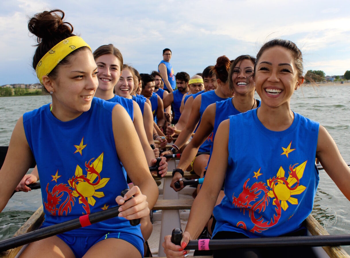 Team rowing on a lake, wearing matching blue shirts with yellow and red dragon design, smiling and holding paddles