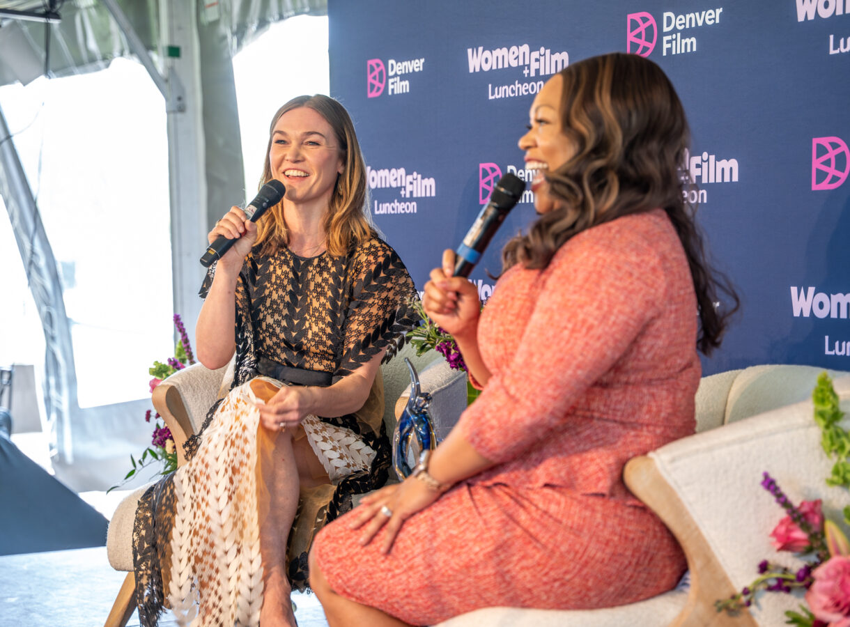 Julia and another woman smiling and speaking into microphones on stage at Women+Film Luncheon, Denver Film backdrop