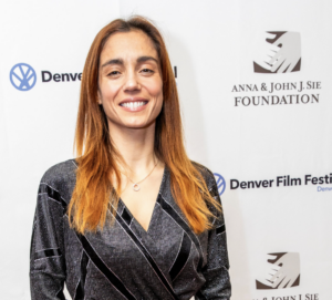 Cristiana Dell'Anna smiling at Denver Film Festival, wearing a gray dress, with event logos in the background