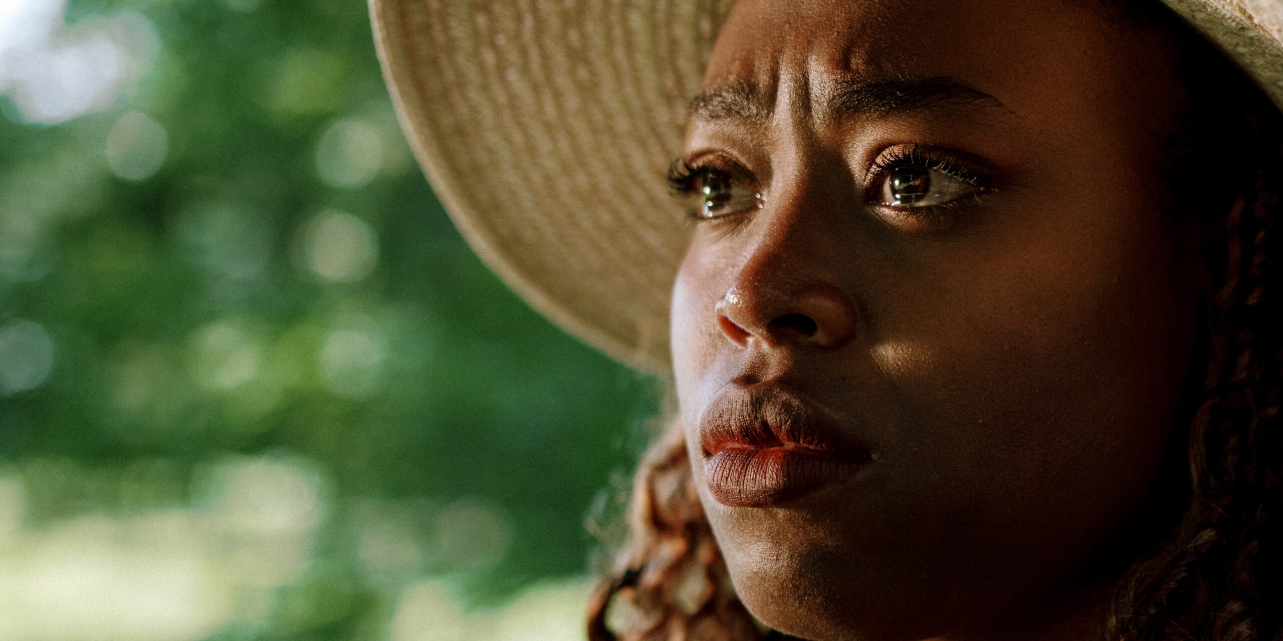 Deja with a serious expression wearing a wide-brimmed hat against a blurred green background
