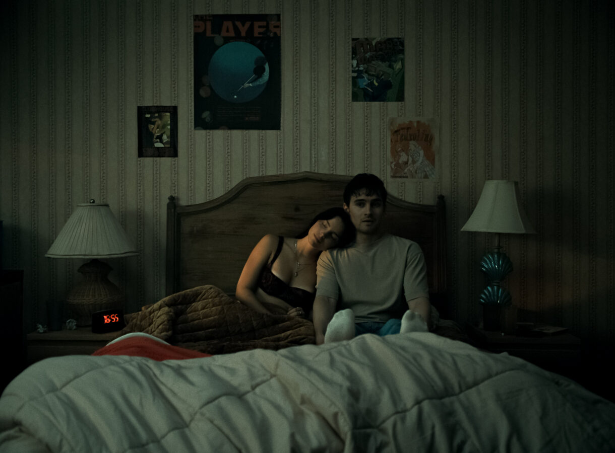 Couple sitting on bed with woman resting on man's shoulder, surrounded by wall posters, clock showing 16:55