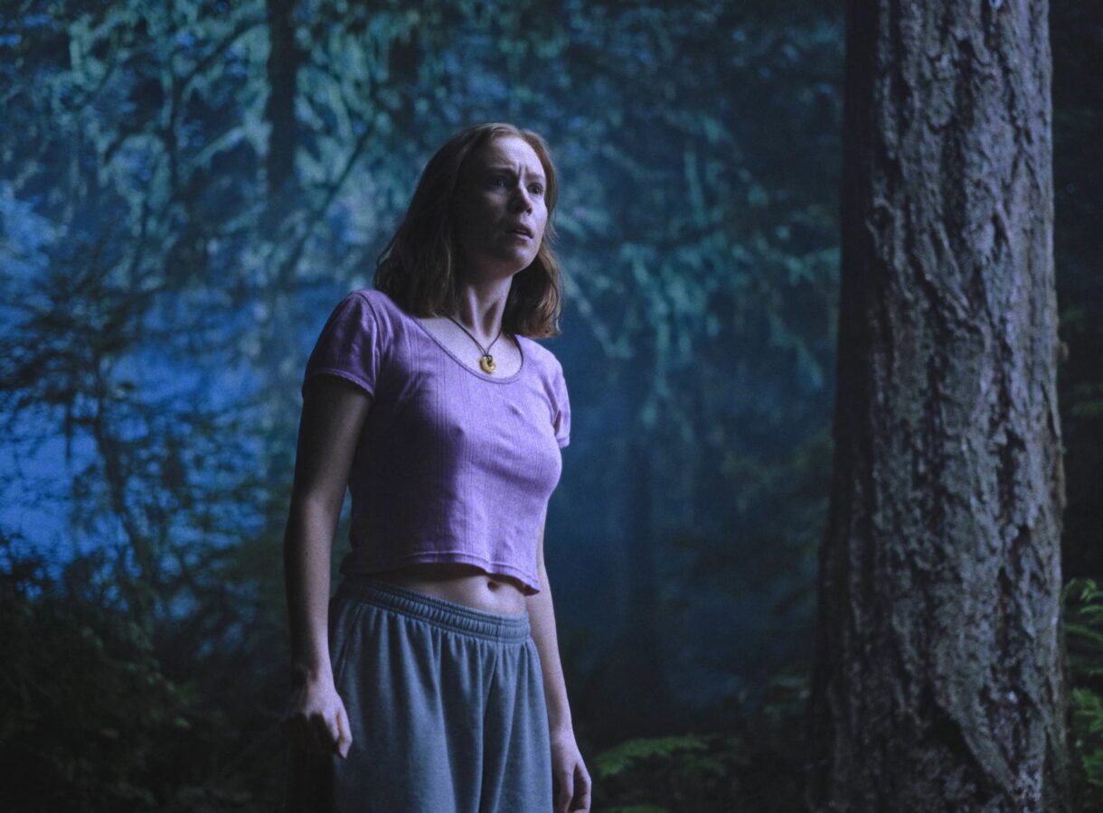 A woman stands in a shadowy forest, looking concerned