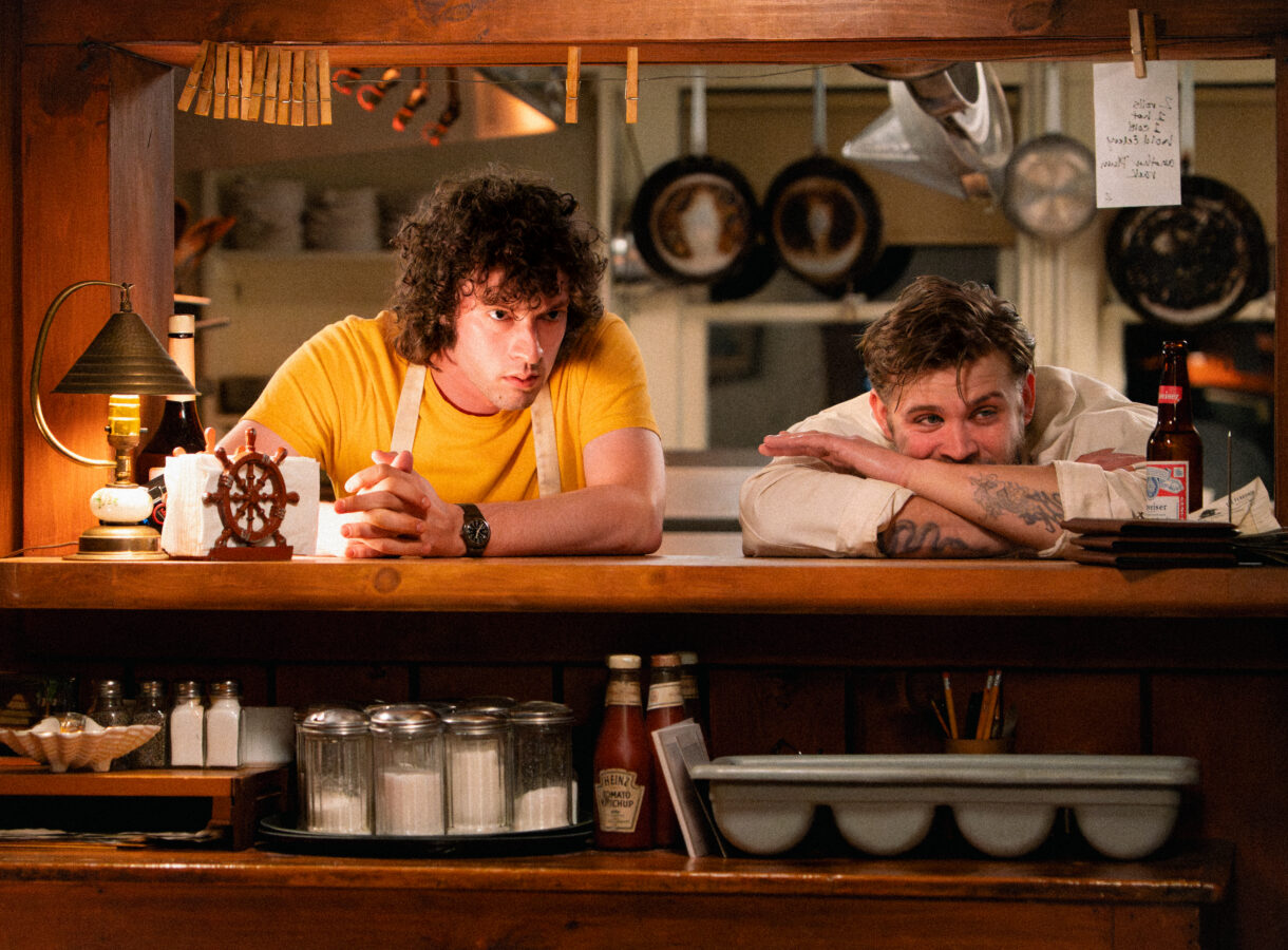 Two men behind a diner counter, one leaning on his arms, the other sitting pensively in a yellow shirt